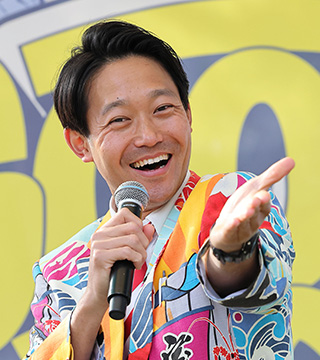 大漁旗をイメージさせる鮮やかなスーツで、マイク片手にポーズをとるハッピーマックスみしまさん